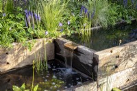 Water rill made from reclaimed timber sleepers with a metal water spout - mixed perennial planting Geranium 'Johnston Blue'