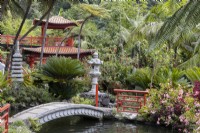 An arched stone bridge leads over a pool, with stone pagoda statues, Oriental style, red balustrades and a large, ornate pagoda in the background. Flowering shrubs are in the foreground and a variety of tropical foliage surrounds the statues and pagoda. Monte Palace Gardens, Madeira. August. Summer