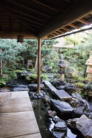 Covered veranda at the back of the house with view into the garden. Rocks placed as stepping stones across water with two stone lanterns or Ishidoro. Straw wrapped fragile stone ornaments as protection against frost.