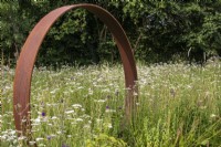 A Corten steel moon gate stands in a perennial meadow with Achillea millefolium.