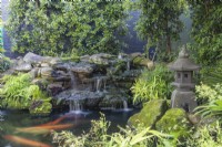 Caption Koi carp pond with a Japanese stone pagoda lantern and Buddha statue near Maiden Hair Fern.
