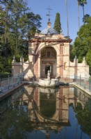 The Cenador del Leon, Lion Pavilion, reflected in a pool surrounded by metal railings and with a  scultpure of a lion within the pool.  Real Alcazar Palace gardens, Seville. Spain. September. 