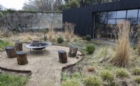 A chipped, circular fire pit area, with large bowled metal fire pit surrounded by tree stump seats. A dark wood clad building is behind with large glass and metal windows and doors. Borders full of plants and grasses are around the fire pit. Winter. Italian Garden at Great Ambrook. 