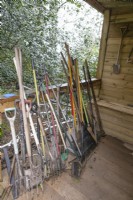 A large selection of gardening tools including spades, forks and rakes on a wooden verandah with various other tools hung on the wall behind. 