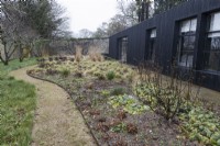 A chipped and curving path leads into the garden on the left, with dark wood clad building on the right. in the centre is a garden full of plants and grasses. Winter. Italian Garden at Great Ambrook. 