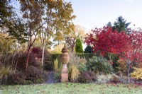 Autumn borders full of trees and shrubs and ornamental grasses around a large terracotta urn.