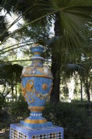 A colourful, glazed urn, on top of a plinth. Various shrubs and trees are in the background.  Parque de Maria Luisa, Seville, Spain. September