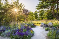 Gravel path leading to seating area with wooden table and two chairs. Planting includes Salvia nemerosa 'Caradonna', Deschampsia cespitosa 'Goldschleier', Eryngium. RHS Iconic Horticultural Hero Garden, Designer: Carol Klein, RHS Hampton Court Palace Garden Festival 2023
