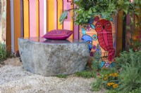 A stone bench with a pink cushion next to a painted old oil drum planted with vegetables, herbs and flowers. The RHS and Eastern Eye Garden of Unity, Designer: Manoj Malde.