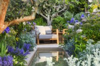 View across the water feature to secluded seating area with armchair  surrounded by Nepeta, Artemisia and architectural fig Ficus carica. Hamptons Mediterranean Garden, Designer: Filippo Dester Garden Club London, RHS Chelsea Flower Show 2023