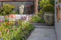 A colourful perennial bed along modern paving slabs and next to a large ceramic vase planted with a yew ball in a contemporary garden. Garden: Looms  and  Blooms, Designer: Kevin Dennis, Bord Bia Bloom 2023