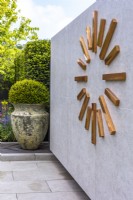 Giant modern copper wall clock decorates a tiled wall. Large ceramic vase with yew ball next to wall.