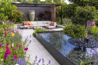 A view of the modern garden with a seating area under a steel pergola, colourful flower beds and a water feature with a trimmed yew tree in the middle. Designer: Kevin Dennis, Bord Bia Bloom 2023 