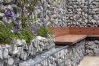 A seating area using reclaimed wood against a boundary wall made of gabions filled with stone offcuts. Geranium 'Johnston Blue' is planted into the gabions - - Caroline and Peter Clayton - Get Started Gardens - Nurturing Nature in the City, RHS Hampton Court Palace Garden Festival.