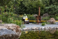 A pool with pebble edge and large boulder leading to a seating area with outdoor wood-burning stove - Hurtigruten: The Relation-Ship Garden - designer Max Parker-Smith - RHS Hampton Court Flower Palace Garden Festival 2023.