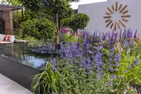 A perennial bed planted with Nepeta and Salvia nemerosa surrounds a modern water feature with a clipped yew tree in the middle. A giant copper wall clock in the background. Designer: Kevin Dennis, Bord Bia Bloom 2023
