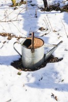 Traditional metal watering can on ground surrounded by snow. 