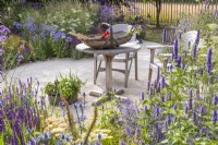 Seating area with wooden table and two chairs surrounded by summer flowering perennial including Salvia nemerosa 'Caradonna', Agastache 'Black Adder' and Eryngium. RHS Iconic Horticultural Hero Garden, Designer: Carol Klein, RHS Hampton Court Palace Garden Festival 2023
