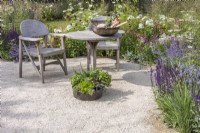 A seating area on a gravel surface with wooden table, two chairs and inset herb planter surrounded by summer border planted with perennials and ornamental grasses. RHS Iconic Horticultural Hero Garden, Designer: Carol Klein, RHS Hampton Court Palace Garden Festival 2023