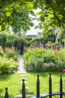View over iron gate along the path of a town garden between flowerbeds filled with Roses, foxgloves, ferns and perennials. Lawn with stepping stones. June.