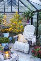 Recycled plastic chair with blankets and cushions, wooden crate and plants adorned with fairy lights inside a greenhouse