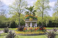 Trachycarpus underplanted with a colourful spring display of red and yellow tulips in a large stone bowl supported by four winged lions, known as the Griffin Tazza (or Lion Vase) in Avenue Gardens in The Regent's Park, London, UK 