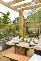 Wooden pergola with a protective perspex roof with a set table in a woodland-inspired garden surrounded by a wooden planks fence. June
Designer: Mary Anne Farenden. Bord Bia Bloom, Super Garden, Dublin, Ireland.