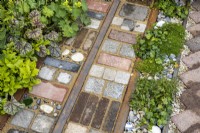 Varied materials: reclaimed cobbles, tiles, bricks, rusted rails and stones used as garden mosaic paving between strip beds planted with Heuchera, Hedera helix and Alchemilla mollis. Designer: Nicola Haines, Citroen Power of One at Bord Bia Bloom Dublin 2023.