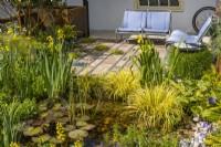 Urban front garden with pond and seating area. Recycled seats are from an old Citroen car model. Mosaic terrace surface made of various reclaimed natural materials. Pond with Nymphaea surrounded by Acorus gramineus 'Tail' and Iris pseudacorus. 
