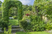 Carpinus betulus - hornbeam arch over low stone steps between lawns at different levels. White painted timber and wrought iron garden bench. Beds and borders with shrubs and perennials. June.