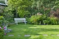 View of a corner of a secluded town garden with white painted timber and wrought iron garden bench on lawn with stepping stones. Informal flowerbeds with peonies and climbing roses. June.