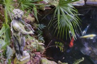 Statue of Pan with flutes, and Trachycarpus fortunei palm leaves above a still Koi Carp pond